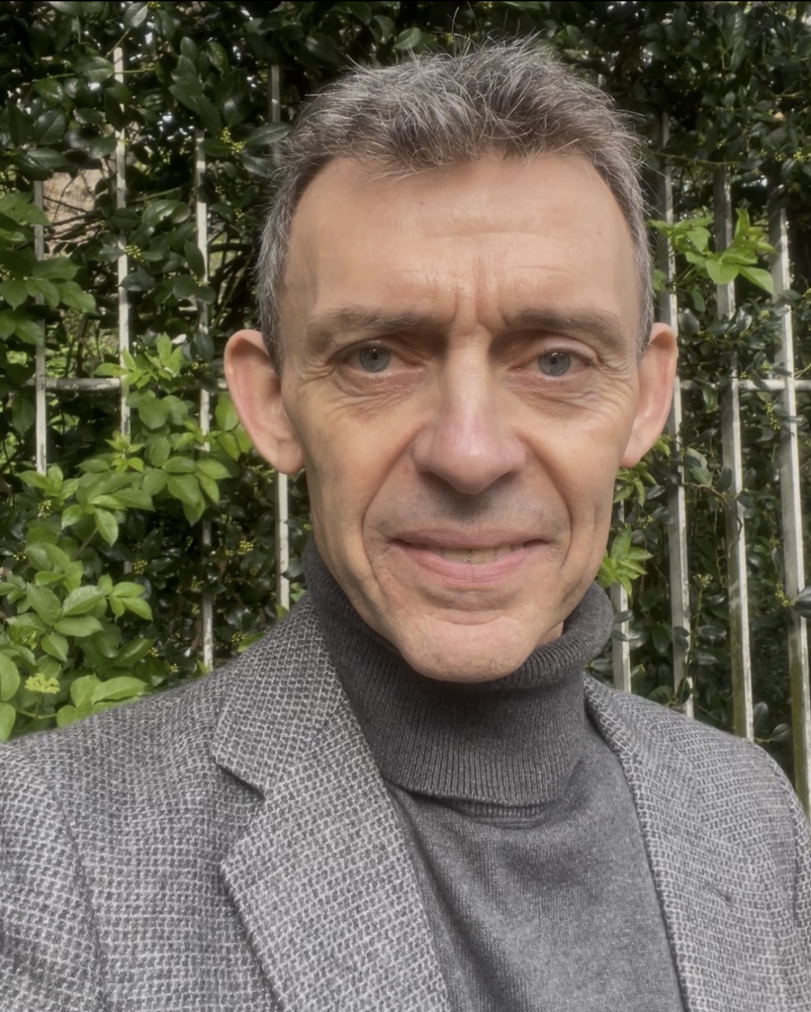 Portrait de Frédéric Rouelle, psychologue, portant un col roulé gris et une veste grise, devant un fond végétal extérieur.